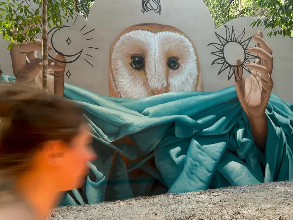 Public art integrated into the jungle at Holistika Tulum: a visitor walks past a mural of a barn owl holding sun and moon symbols.