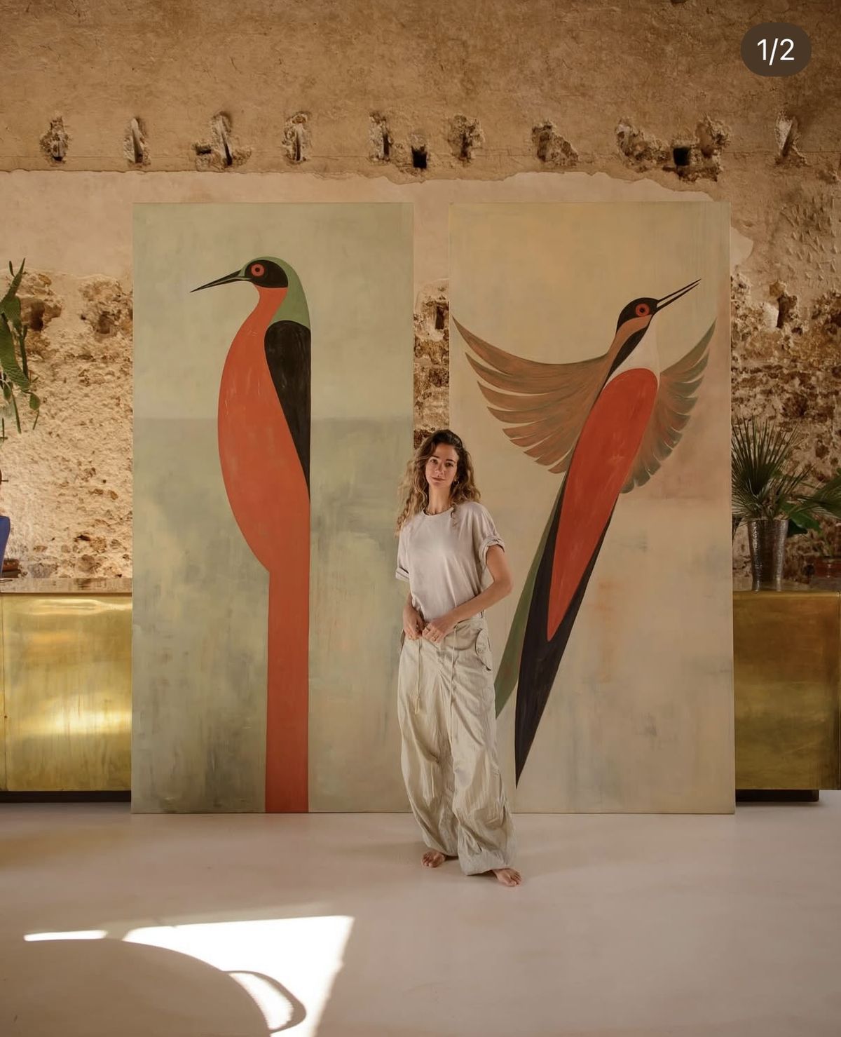 A woman standing between two tall paintings of stylised tropical birds in coral, green and charcoal tones against a weathered stone wall.