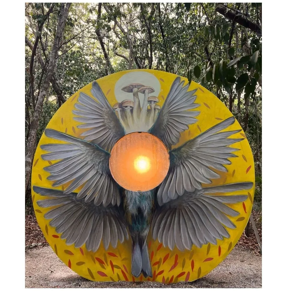 Circular yellow panel of outstretched bird wings around a glowing amber light, standing among the trees at Holistika Art Walk in Tulum.