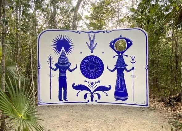 Cobalt blue mural of two ceremonial figures flanking a spiral, painted on a white panel standing between palms in the jungle at Holistika.