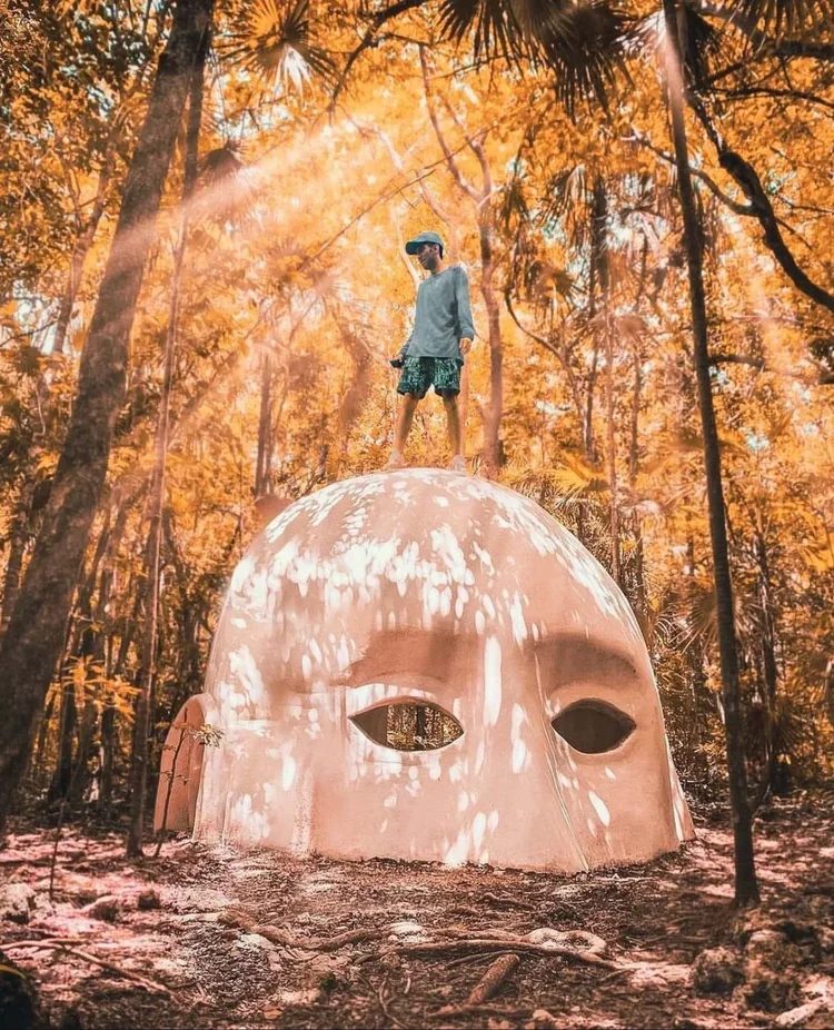Holistika Art Walk mural surrounded by jungle in Tulum, Mexico, with a visitor standing on a large face shaped dome sculpture.